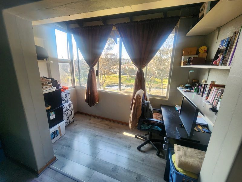 A compact home office space featuring laminate flooring, built-in shelving, and large windows providing natural light and exterior views.