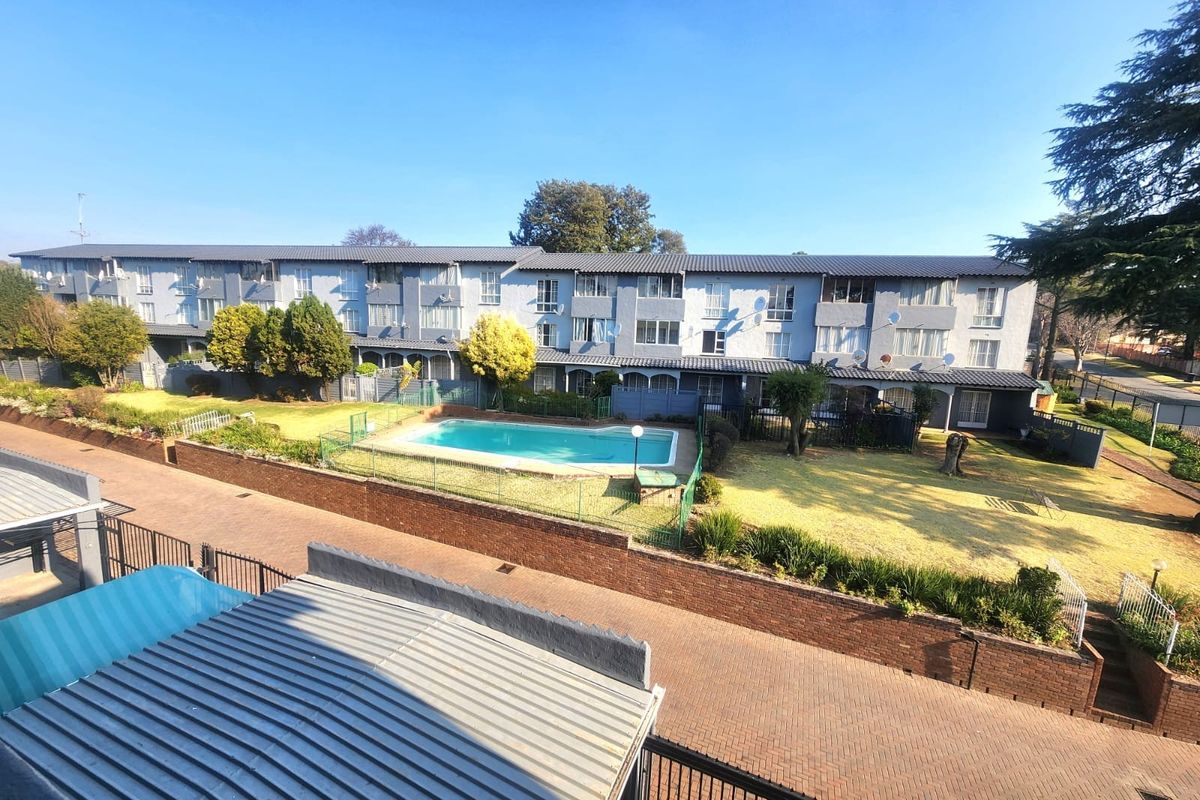 Elevated view of the apartment complex featuring a communal swimming pool, landscaped garden areas, paved driveways, and secure perimeter fencing.