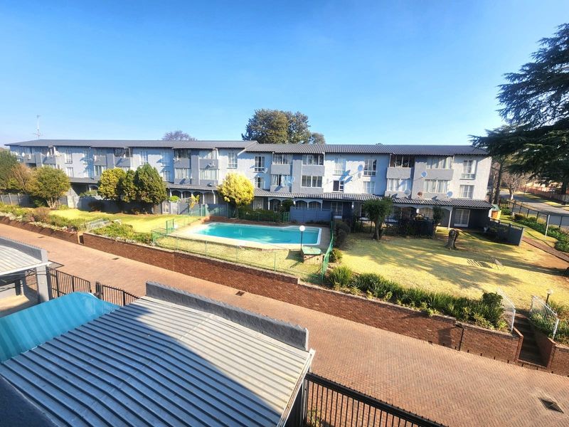 Elevated view of the apartment complex featuring a communal swimming pool, landscaped garden areas, paved driveways, and secure perimeter fencing.