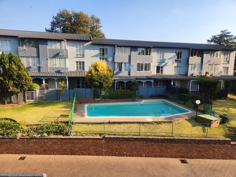 Exterior view of an apartment complex featuring a communal swimming pool, landscaped garden area, and paved driveway access.