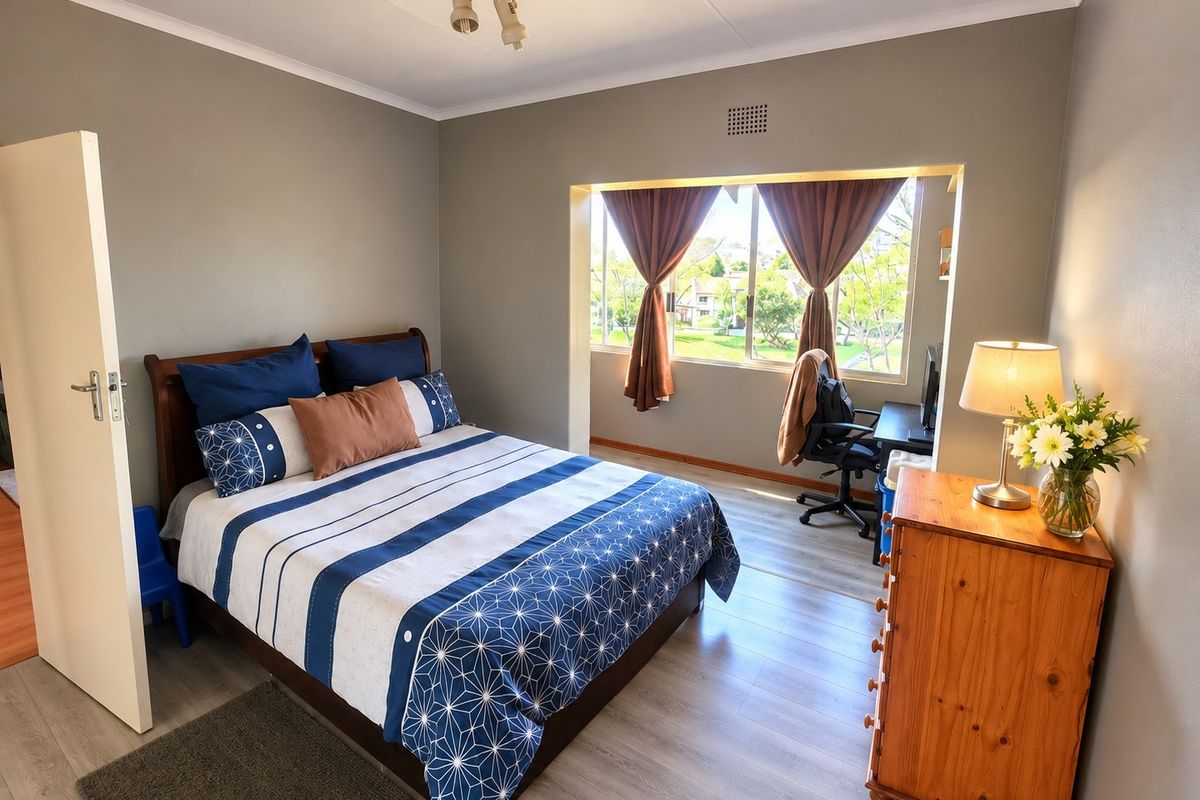 A well-lit bedroom featuring laminate flooring, a large window with natural light, and a neutral wall finish.