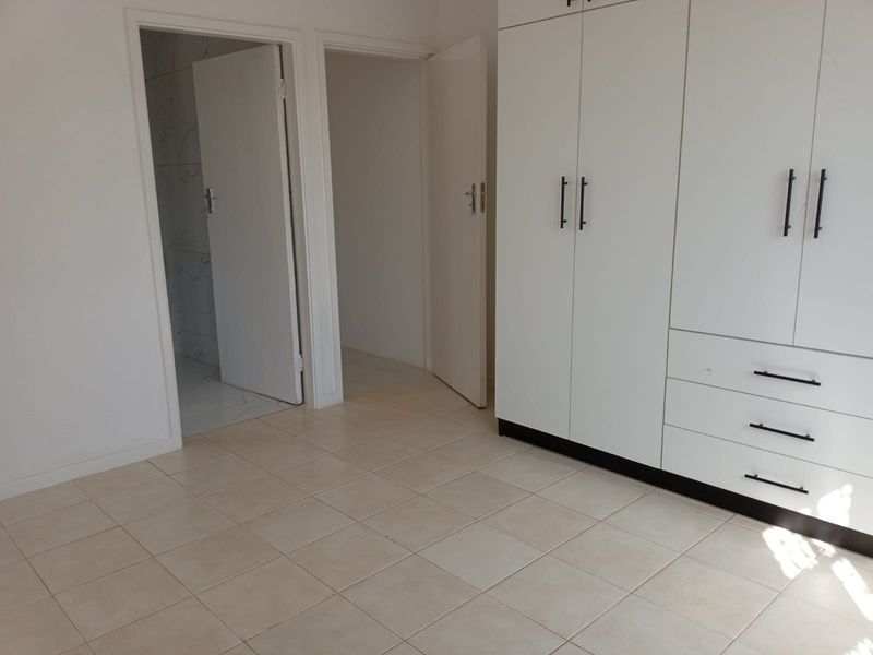 A clean, empty bedroom featuring neutral tiled flooring and white built-in cupboards, with direct access to an adjacent bathroom and hallway.