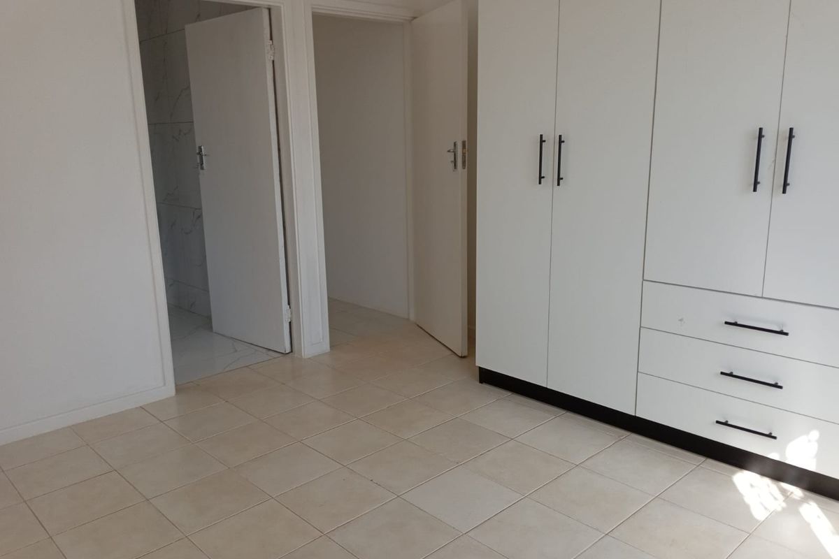 A clean, empty bedroom featuring neutral tiled flooring and white built-in cupboards, with direct access to an adjacent bathroom and hallway.