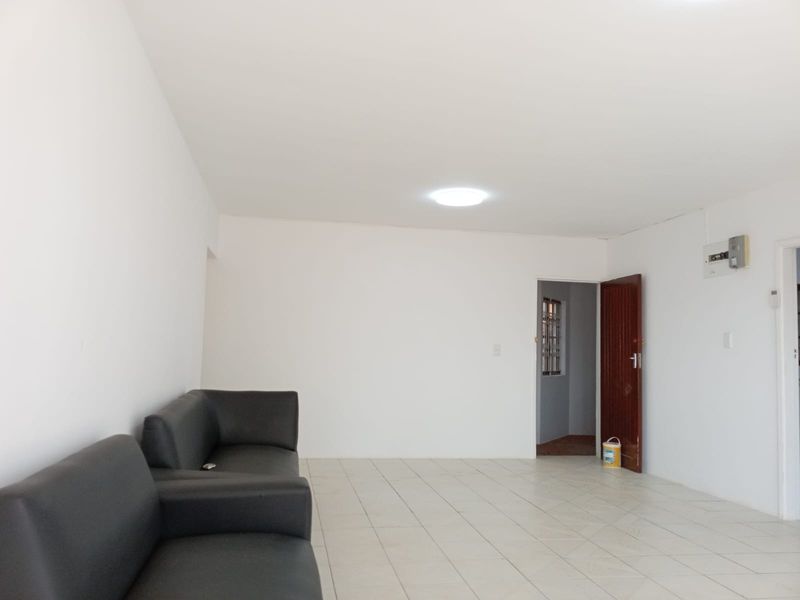 A bright, open-plan living area featuring neutral tiled flooring, white walls, and a functional layout with access to an adjacent room.