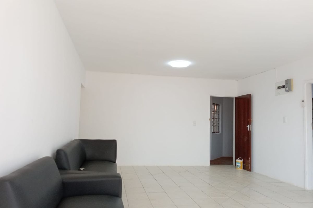 A bright, open-plan living area featuring neutral tiled flooring, white walls, and a functional layout with access to an adjacent room.