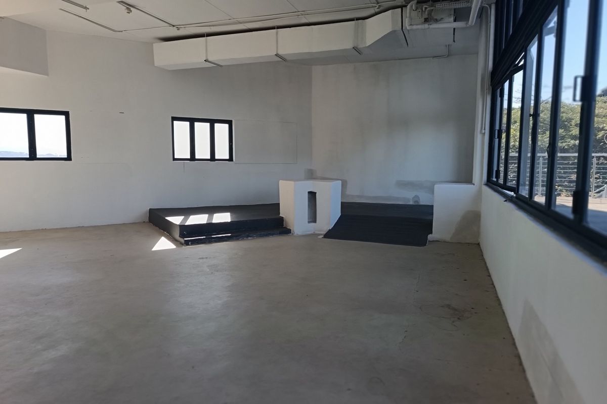 An empty, open-plan commercial space featuring exposed ductwork, concrete flooring, and a raised platform area with large steel-framed windows.