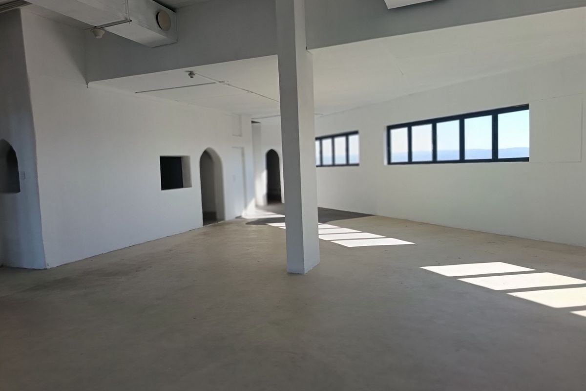 A spacious, empty open-plan restaurant area featuring white walls, exposed structural columns, arched doorways, and large horizontal windows providing natural light.
