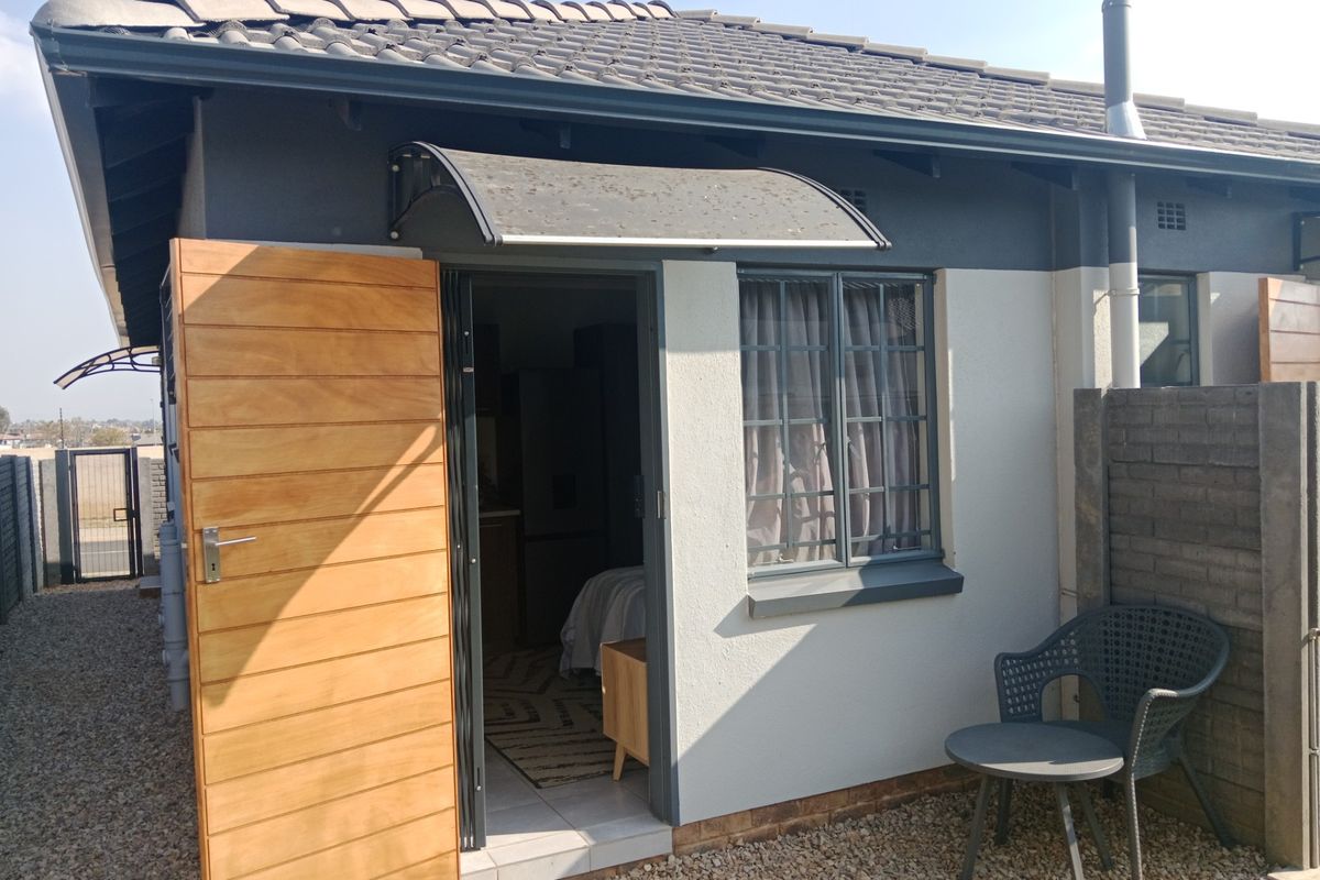 Rear exterior view of the property featuring a modern timber door, protective awning, and secure window bars, set against a low-maintenance gravel courtyard.