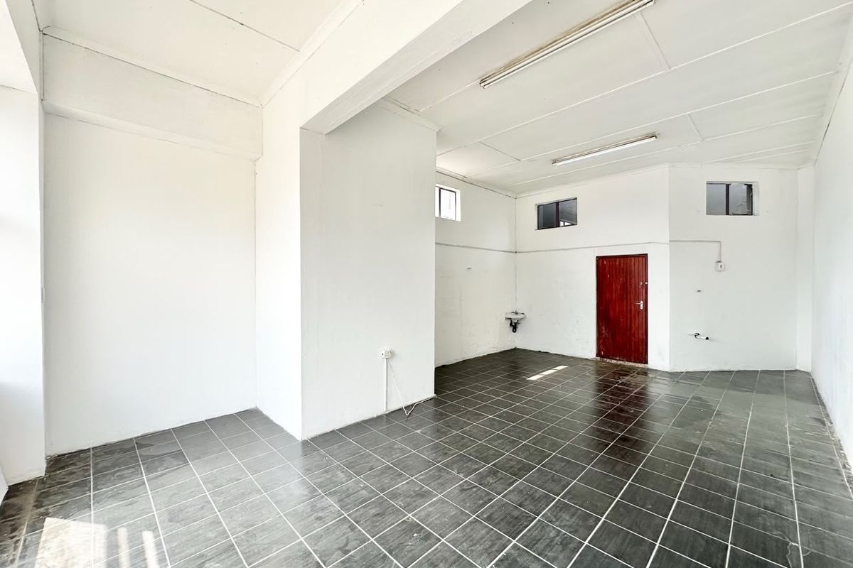 An empty, open-plan commercial space featuring dark tiled flooring, fluorescent lighting, and a small wall-mounted basin, suitable for various business or retail applications.