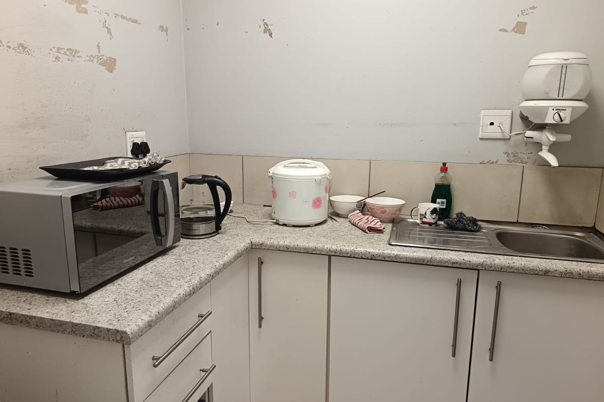 A compact kitchen area featuring white cabinetry, a stainless steel sink, and a wall-mounted water heater, currently requiring wall surface repairs and cosmetic updates.
