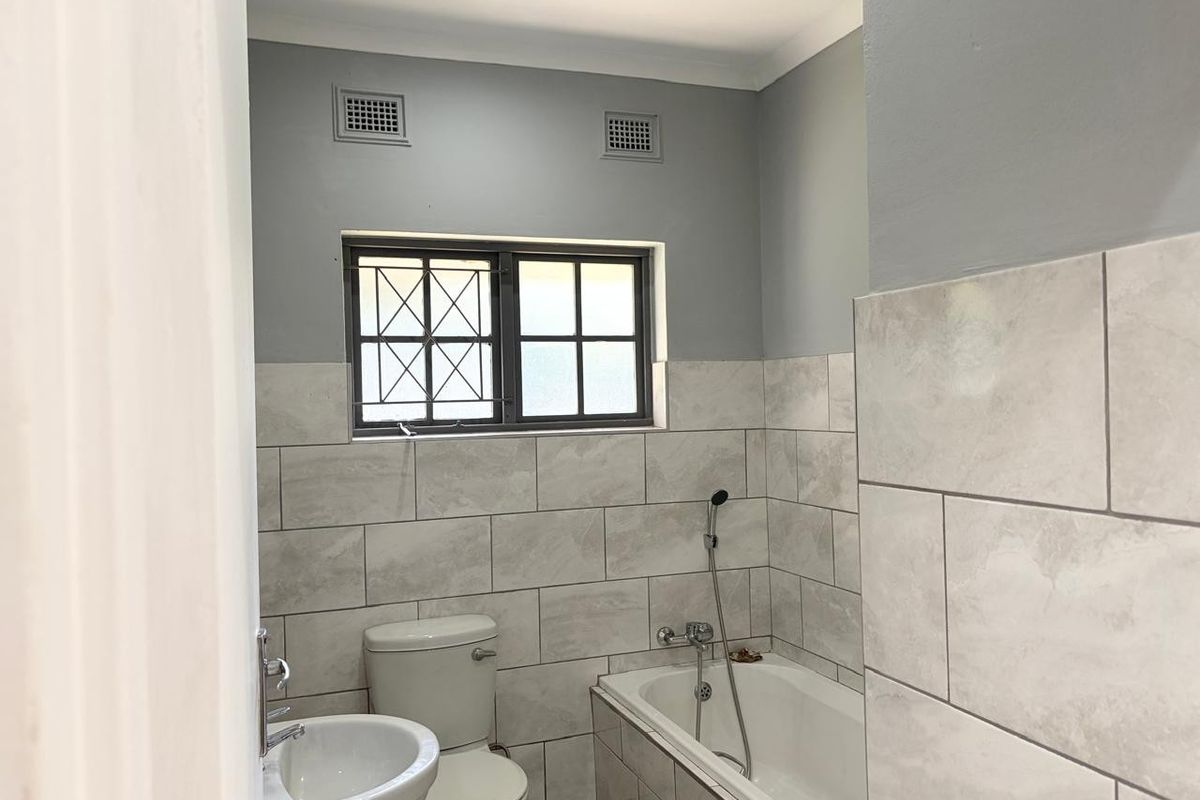 A clean, modern bathroom featuring neutral wall tiles, a pedestal basin, a toilet, and a bathtub with a handheld shower attachment.