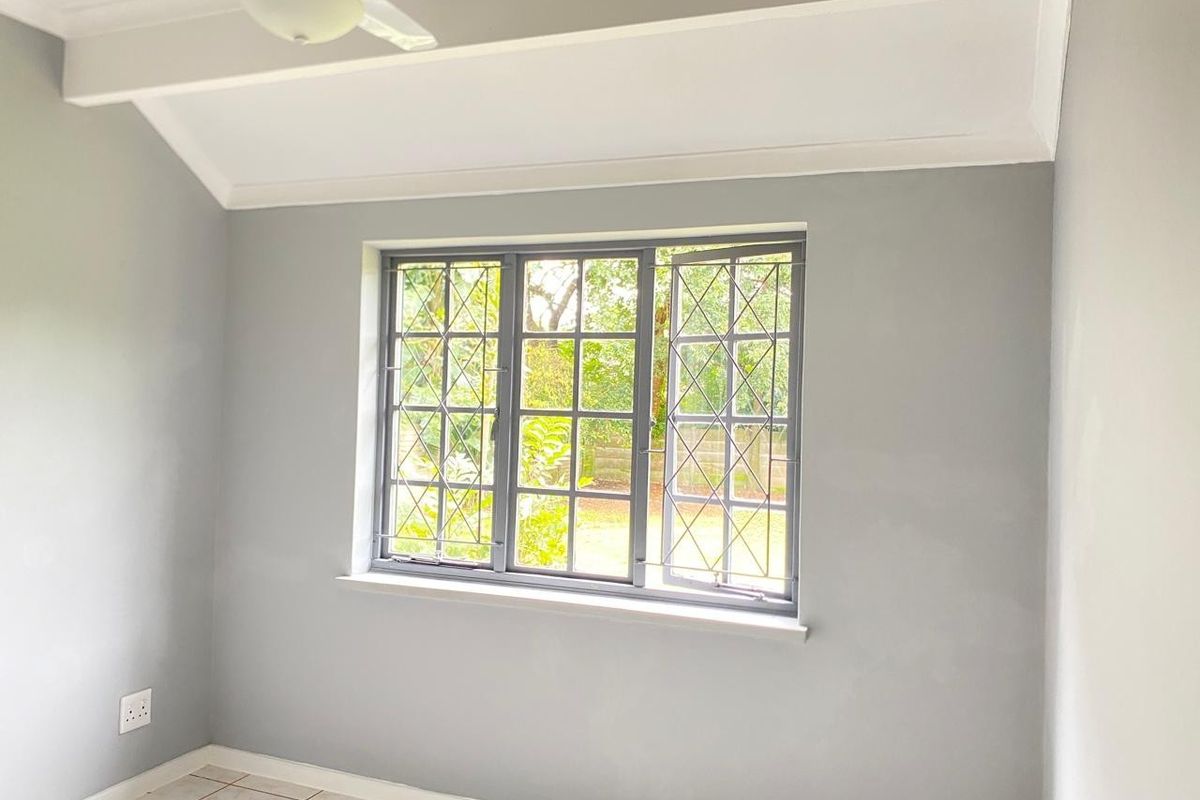 A bright, empty bedroom featuring neutral grey walls, tiled flooring, a ceiling fan, and a classic leaded glass window overlooking the garden.