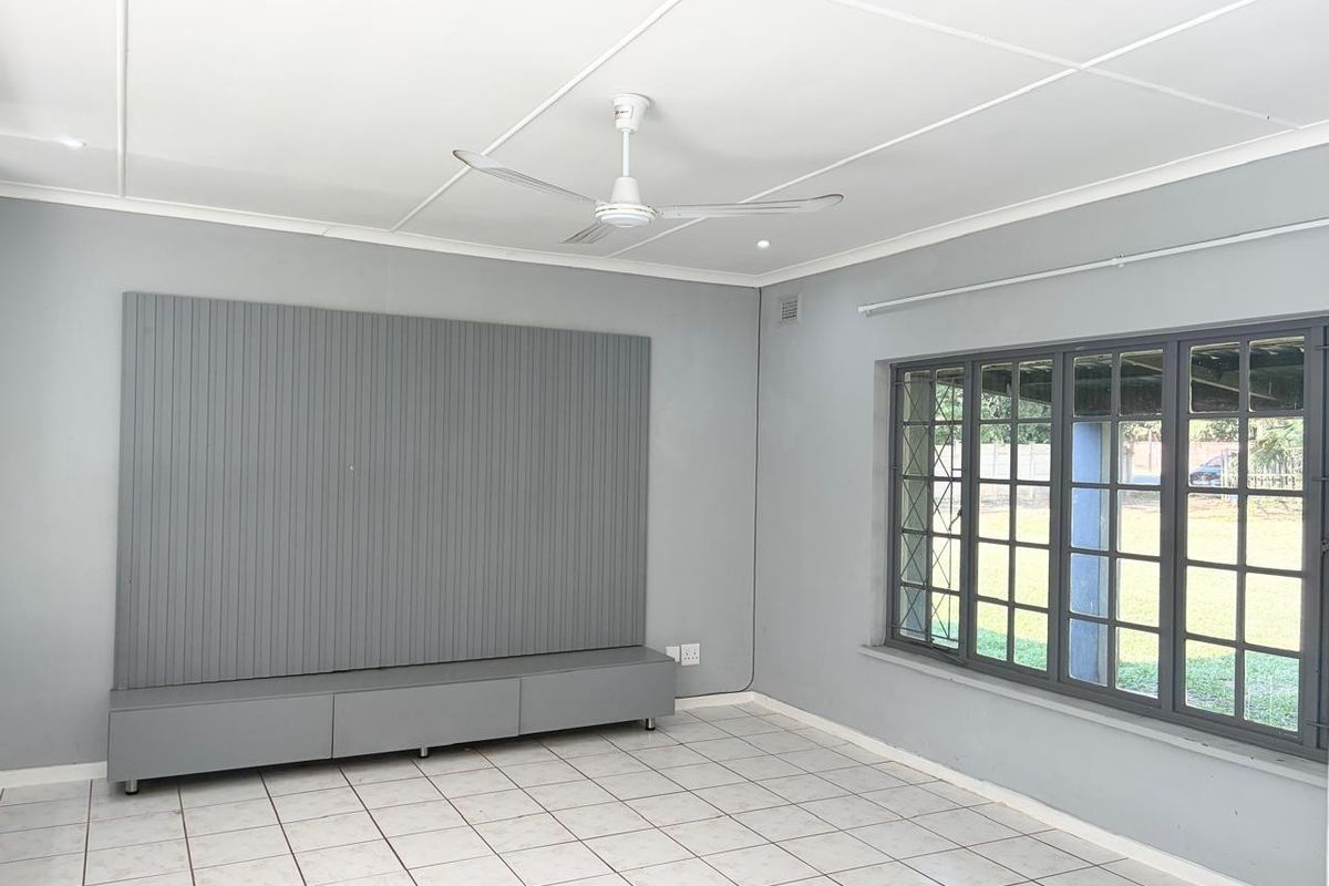 A bright, empty living room featuring neutral grey walls, tiled flooring, a ceiling fan, and a built-in media console with decorative wall paneling.