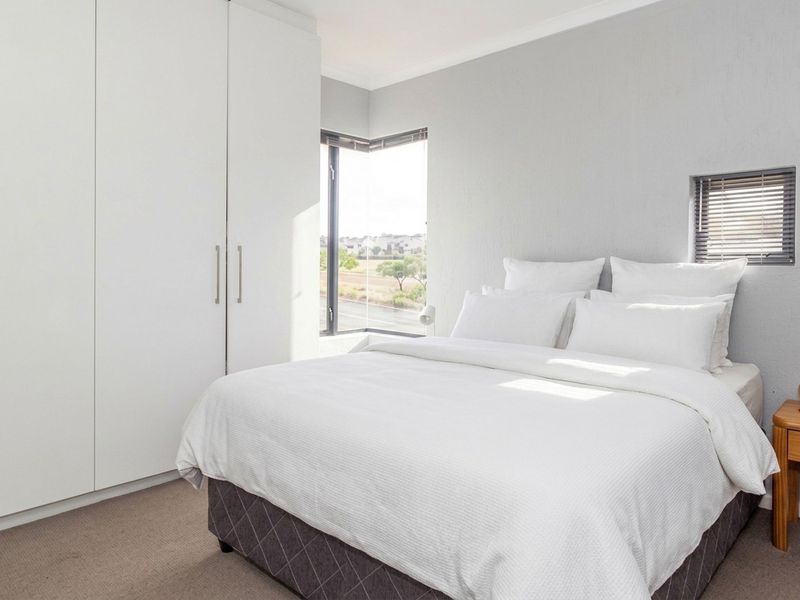 A bright, modern bedroom featuring built-in cupboards, neutral carpeting, and a corner window with venetian blinds offering natural light.
