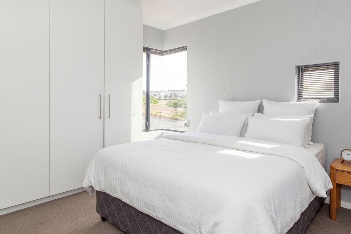 A bright, modern bedroom featuring built-in cupboards, neutral carpeting, and a corner window with venetian blinds offering natural light.