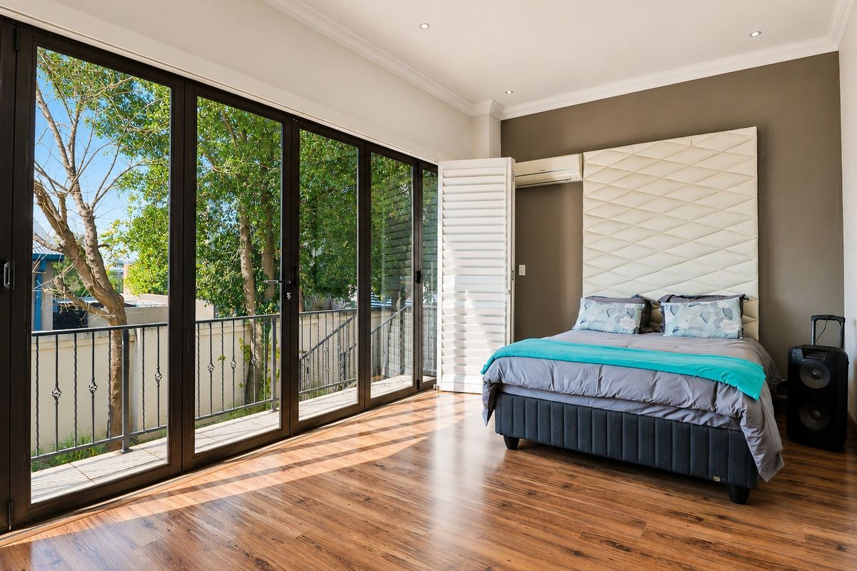 Spacious bedroom featuring warm laminate flooring, a custom upholstered headboard, and large sliding glass doors opening onto a private balcony with garden views.