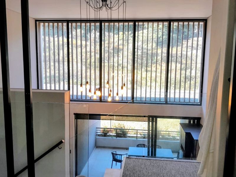 Modern double-volume living space featuring expansive floor-to-ceiling windows, glass balustrades, and elegant pendant lighting, overlooking a private patio area.