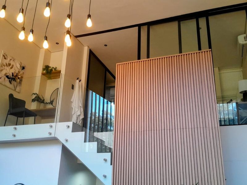 Modern living area featuring a striking slatted timber wall, glass balustrade staircase, and a mezzanine level with contemporary lighting and high ceilings.