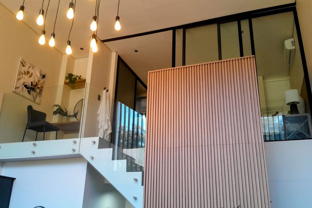 Modern living area featuring a striking slatted timber wall, glass balustrade staircase, and a mezzanine level with contemporary lighting and high ceilings.