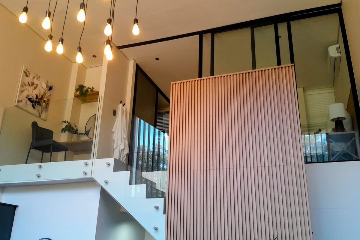 Modern living area featuring a striking slatted timber feature wall, glass balustrade staircase, and contemporary black metal partitions with high-ceiling pendant lighting.