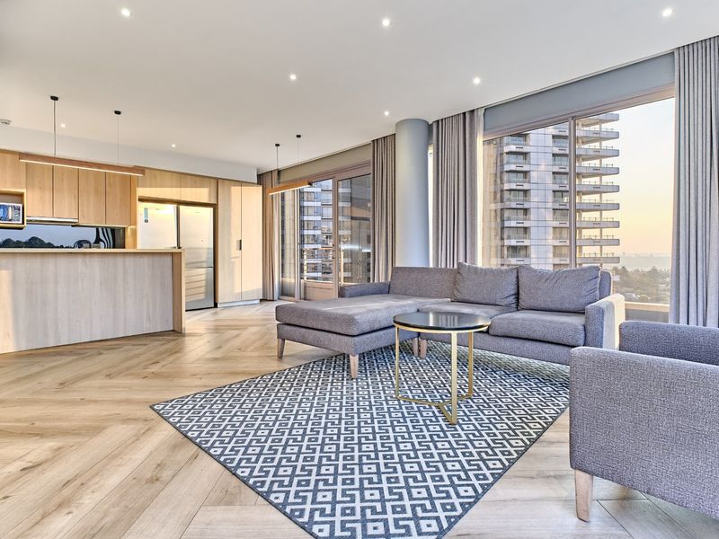 Modern open-plan living area featuring herringbone timber flooring, sleek built-in kitchen cabinetry, and expansive floor-to-ceiling windows with city views.