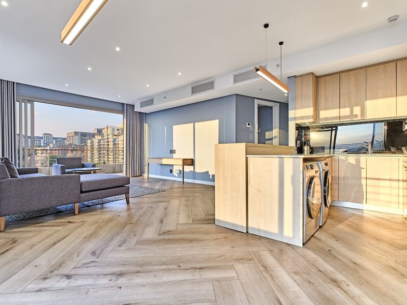 Modern open-plan living area featuring herringbone timber flooring, light wood cabinetry, integrated laundry appliances, and large sliding doors leading to a balcony.
