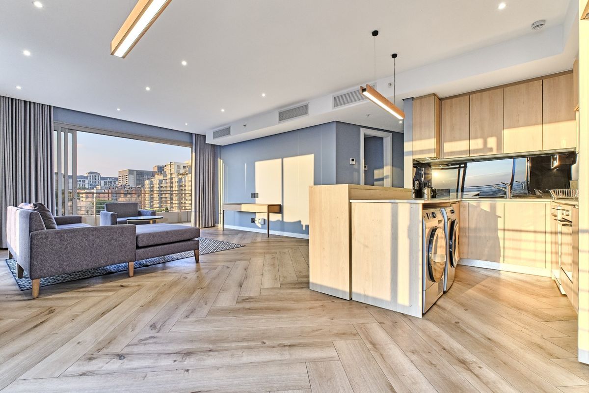 Modern open-plan living area featuring herringbone timber flooring, light wood cabinetry, integrated laundry appliances, and large sliding doors leading to a balcony.