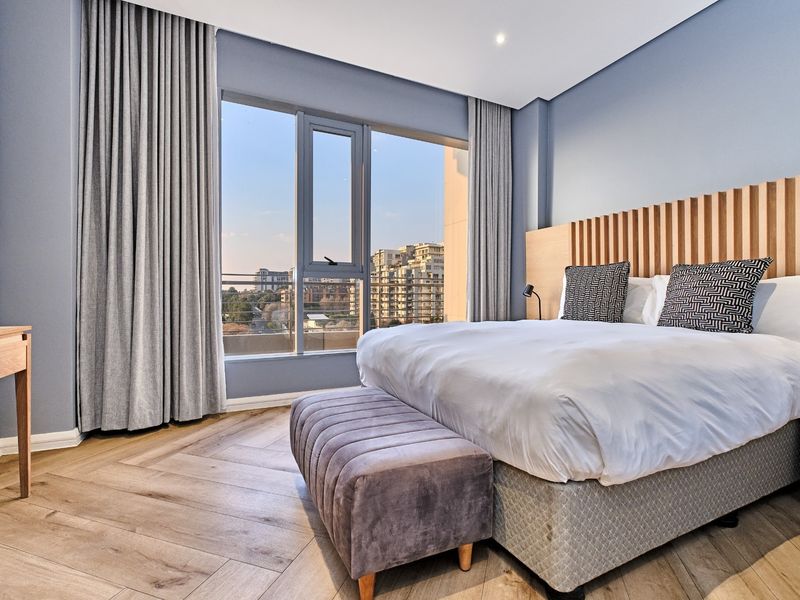 Modern bedroom featuring herringbone wood flooring, a custom timber headboard, and large windows offering urban views with integrated curtain tracks.