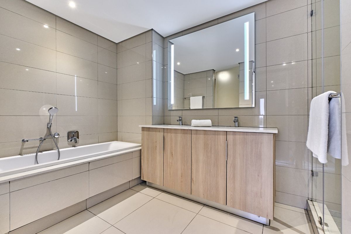 Modern bathroom featuring a double vanity with wood-finish cabinetry, a built-in bathtub, and a glass-enclosed shower with neutral floor-to-ceiling tiling.