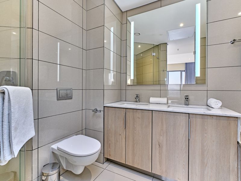 Modern bathroom featuring a double vanity with stone countertop, wall-mounted toilet, glass shower enclosure, and elegant floor-to-ceiling tiling.