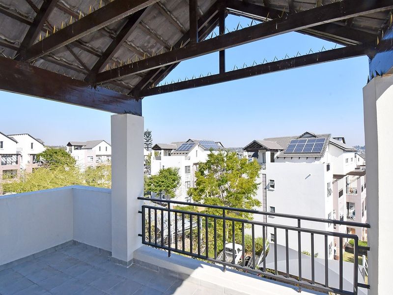 A spacious covered balcony featuring tiled flooring, a secure metal balustrade, and exposed timber roof trusses, offering an elevated view of the surrounding apartment complex.