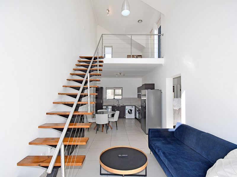 Spacious open-plan living area featuring a modern open-tread staircase, mezzanine level, and a functional kitchen with tiled flooring and high vaulted ceilings.