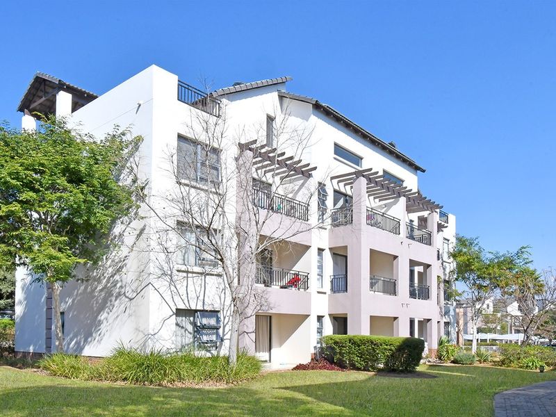 A multi-story apartment building featuring private balconies with pergolas, metal railings, and well-maintained surrounding garden areas with paved walkways.