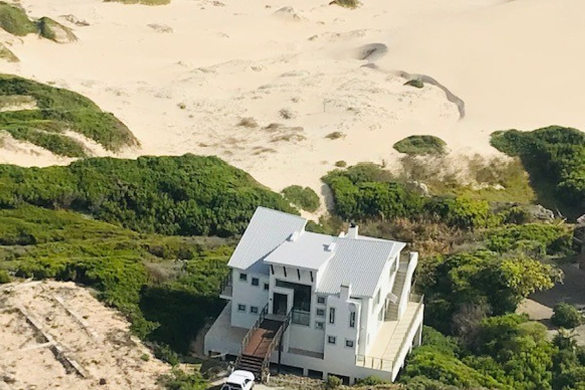 Aerial view of a coastal property featuring a multi-level house with a metal roof, elevated timber entrance walkway, and direct beach access.