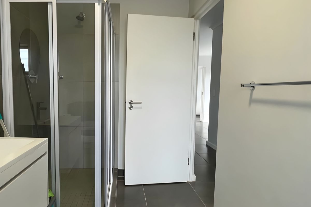 A clean, modern bathroom featuring a glass shower enclosure, vanity cabinet, and neutral tiled flooring, leading into a hallway.