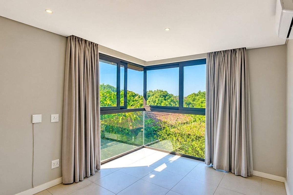 A bright, empty bedroom featuring modern corner windows with a glass balustrade, tiled flooring, and a wall-mounted air conditioning unit.