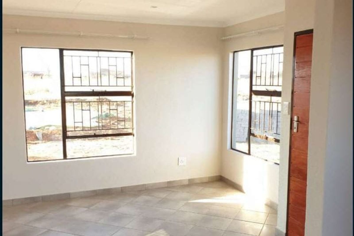 A bright, empty living area featuring neutral tiled flooring, large windows with security bars, and a timber door.