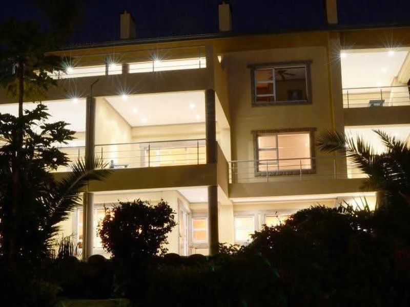 A multi-level rear facade of a modern house at night, featuring illuminated balconies, stainless steel railings, and large glass sliding doors.