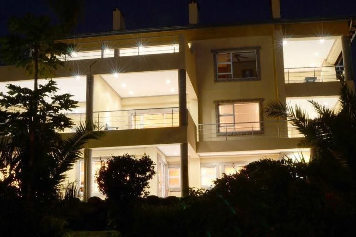 A multi-level rear facade of a modern house at night, featuring illuminated balconies, stainless steel railings, and large glass sliding doors.