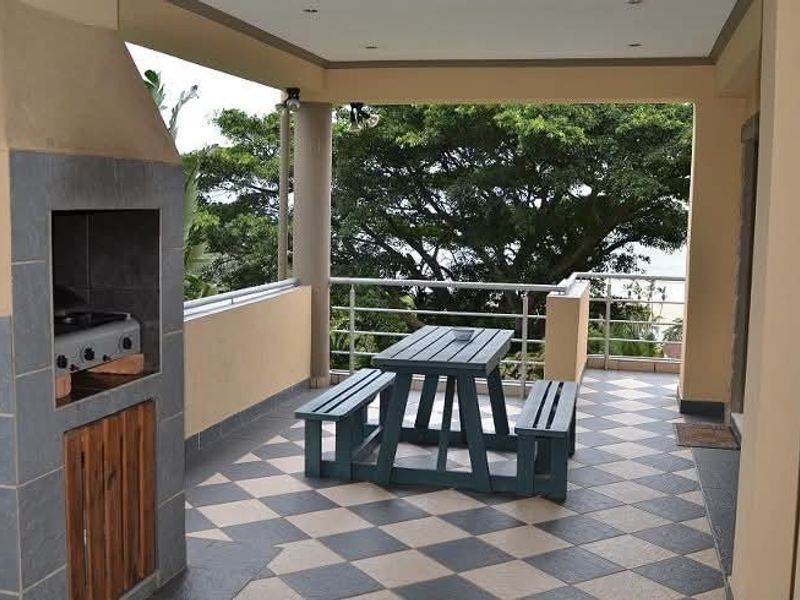 A covered balcony featuring a built-in braai, tiled flooring, and metal railings, offering an outdoor entertainment space with views of surrounding trees.