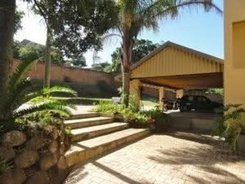 Rear exterior view featuring a paved driveway, covered carport, stone retaining wall, and landscaped garden area with concrete steps.