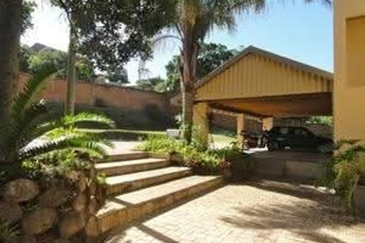 Rear exterior view featuring a paved driveway, covered carport, stone retaining wall, and landscaped garden area with concrete steps.