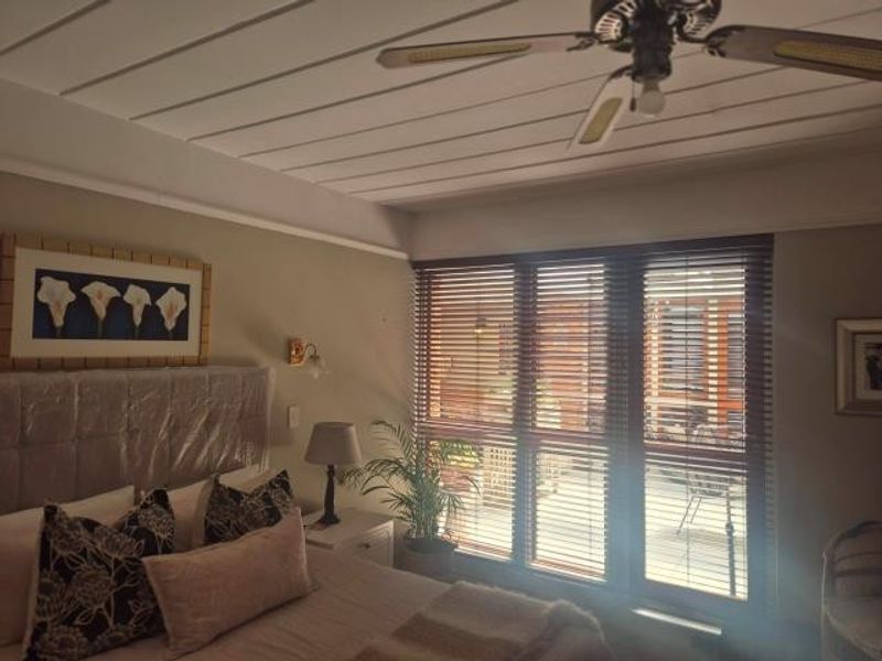 A bedroom featuring a ceiling fan, wall-mounted lighting, and venetian blinds covering a large window, providing natural light and privacy.