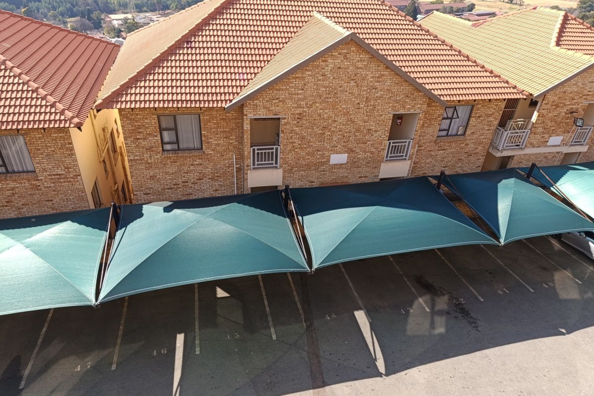 Aerial view of an apartment complex featuring face brick exteriors, tiled roofing, and designated parking bays covered by shade cloth structures.