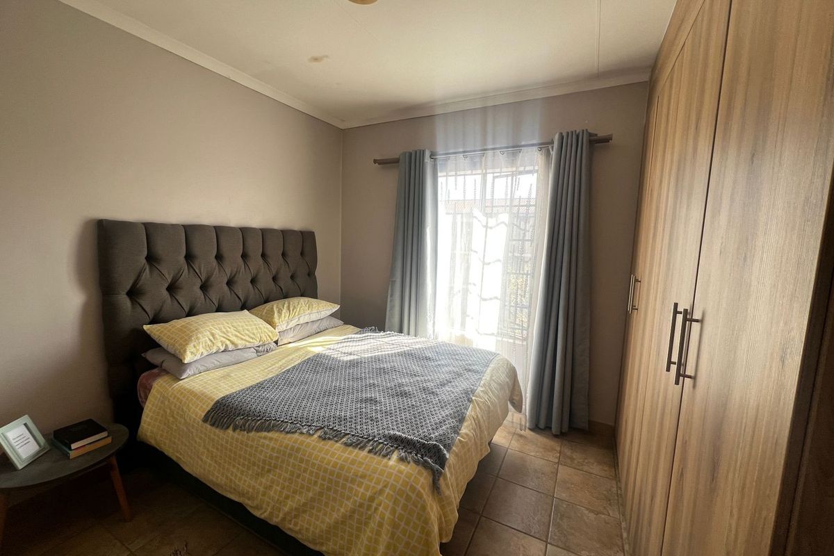 A functional bedroom featuring built-in wooden cupboards, tiled flooring, and natural light through a window with security bars.