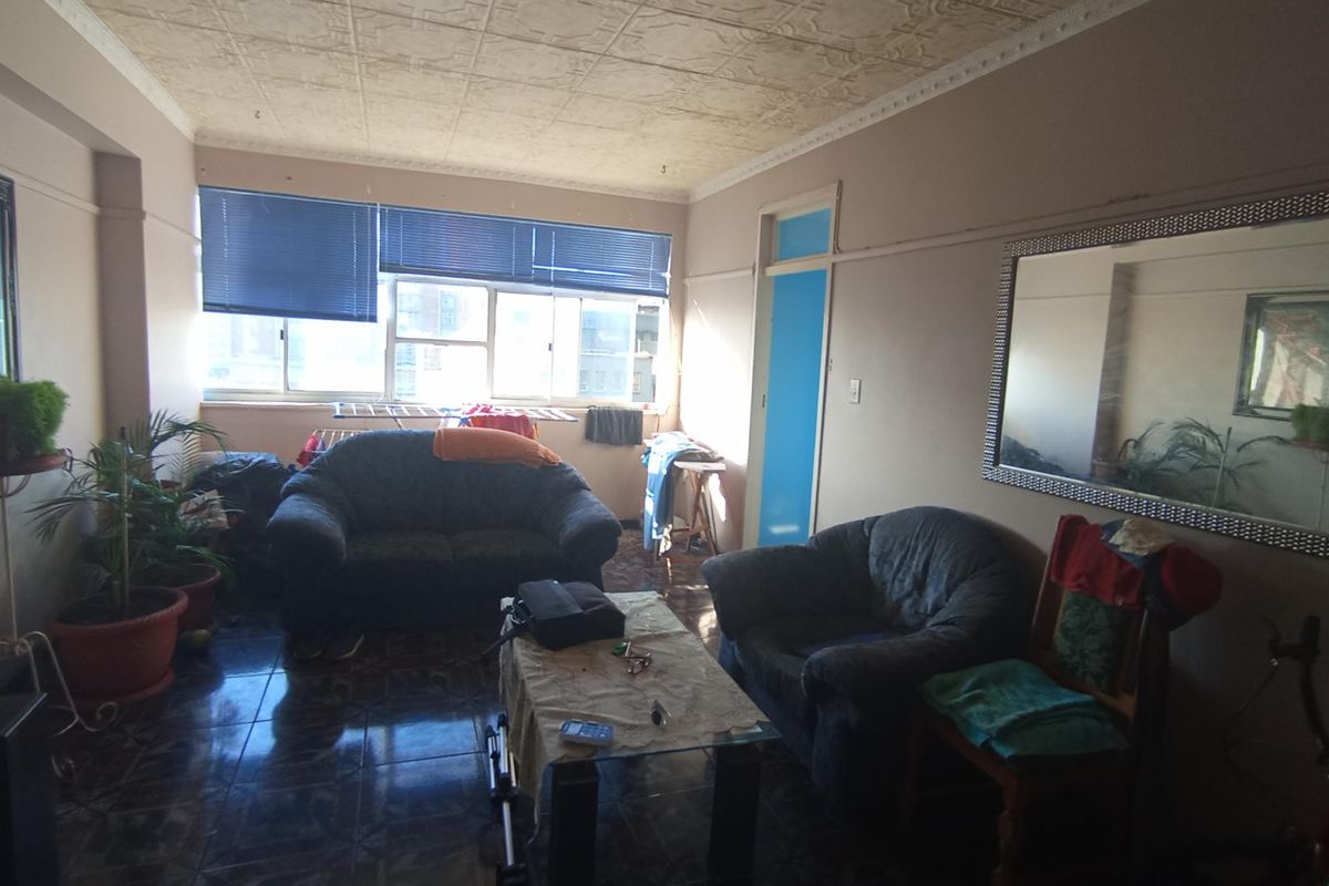 A living room featuring tiled flooring, decorative ceiling tiles, and large windows with venetian blinds, offering a dated but functional space.