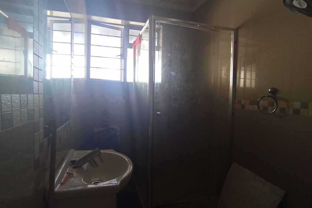 A functional bathroom featuring a shower enclosure, pedestal basin, and tiled walls, benefiting from natural light through a window with security bars.