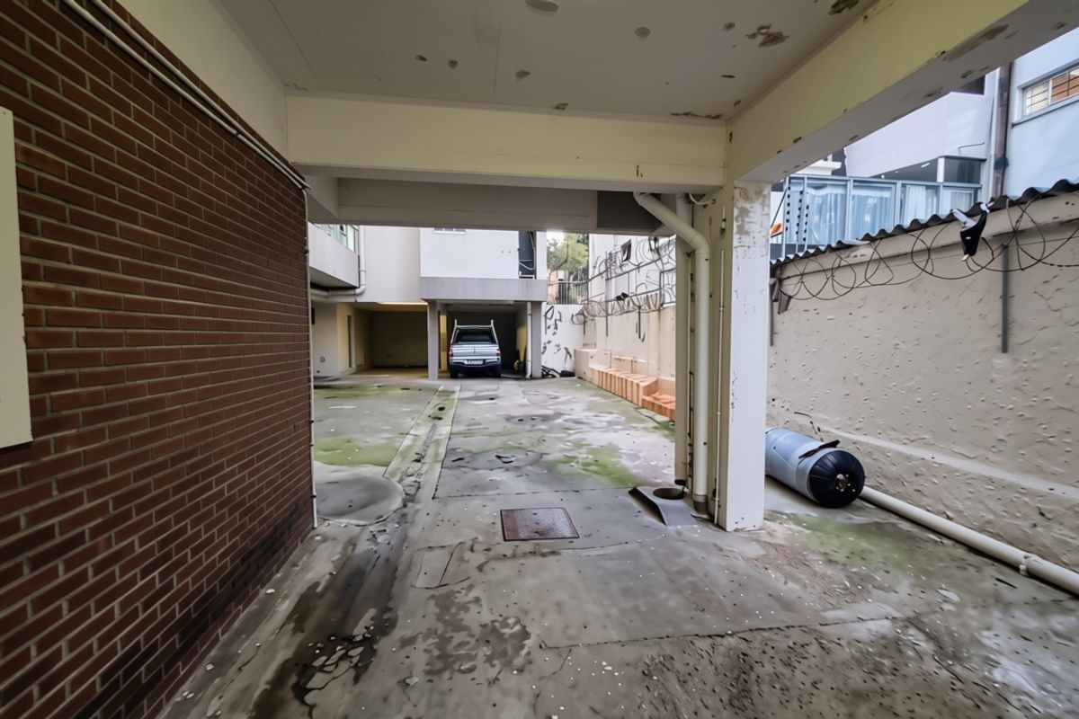 A rear exterior area featuring a covered parking space, brick walling, and concrete paving showing signs of significant wear and maintenance requirements.