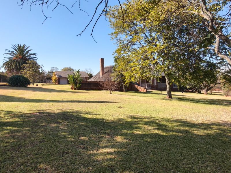 Spacious rear garden view featuring a large lawn, mature trees, and a view of the main residence with a thatched roof and detached garage.