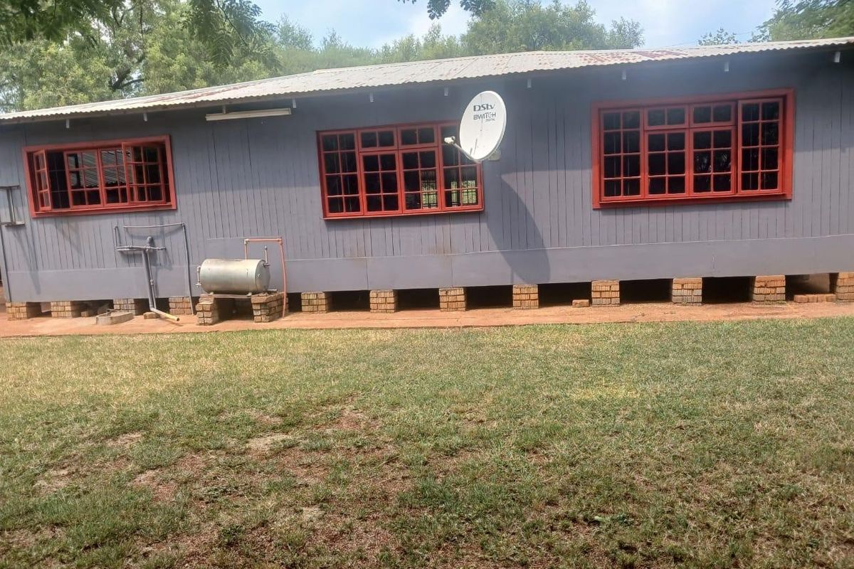 Rear view of a timber-clad small holding structure featuring a corrugated metal roof, brick foundation piers, and red-framed casement windows.